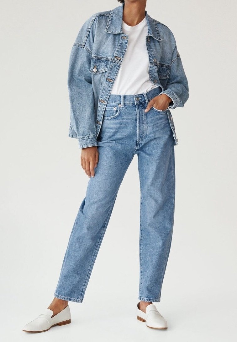 Personne portant une veste en jean bleu clair et un jean avec un t-shirt blanc et des chaussures blanches à enfiler, debout avec une main dans la poche.