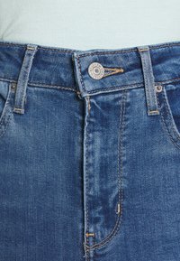 Jeans de mezclilla de color azul, con un botón de metal, costuras de cobre y un diseño de corte recto. Toma de cerca de la zona de la cinturilla.
