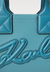 Blue textured leather handbag with dark stitching, featuring a prominent raised logo and structured handles. Smooth finish and detailed accents.