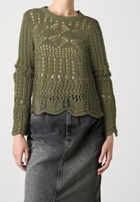 Pull tricoté vert olive avec motif ajouré complexe, manches longues et ourlet festonné, associé à une jupe en denim gris foncé.