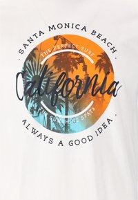Hvid T-shirt med et cirkulært grafisk design med orange og blå gradienter, palmetræer og tekst: "Santa Monica Beach," "California," og mere.