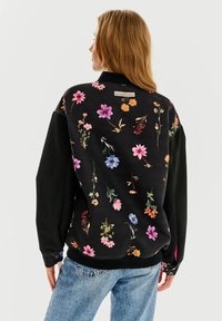 Czarna bluza ze wzorem kwiatowym w różnych kolorach; posiada długie rękawy, ściągacze i metkę z napisem "BUTTERFLY EFFECT" z tyłu.