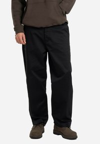 Black, relaxed-fit pants with a smooth texture, featuring a waistband and side pockets. Worn with brown, sturdy shoes.