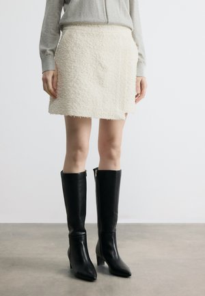 Ženska, oblečena v teksturirano belo mini krilo, sivo bluzo z dolgimi rokavi in črne škornje z visokimi petami do kolen, stoji na sivem tlaku.