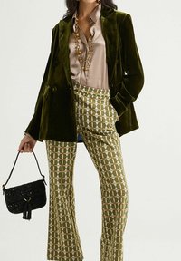 Blazer croisé en velours vert, chemise en satin beige, pantalon évasé à motifs verts et beiges, et sac à paillettes noir avec un pompon.
