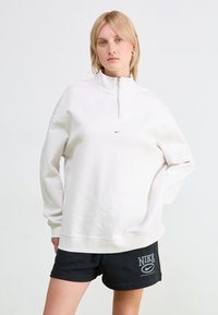 White, textured sweatshirt with a high collar and quarter-zip, featuring a small silver logo on the chest. Paired with black shorts.