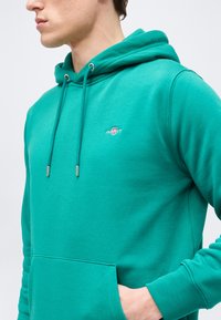 Νεαρός άνδρας που φοράει μια πετρόλ φούτερ με κουκούλα, μπροστινή τσέπη και μικρό κεντημένο λευκό και κόκκινο λογότυπο στο στήθος.