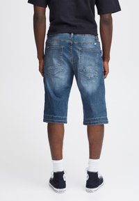 Shorts en denim de bleu moyen, avec une coupe décontractée, longueur genou, couture visible et deux poches arrière. Portés avec des chaussettes blanches et des baskets noires.
