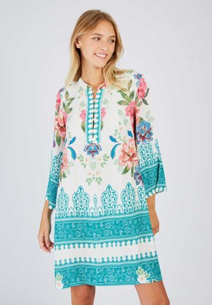 Young woman smiling, wearing a knee-length white dress with colorful floral and turquoise ornate patterns on sleeves and hem.