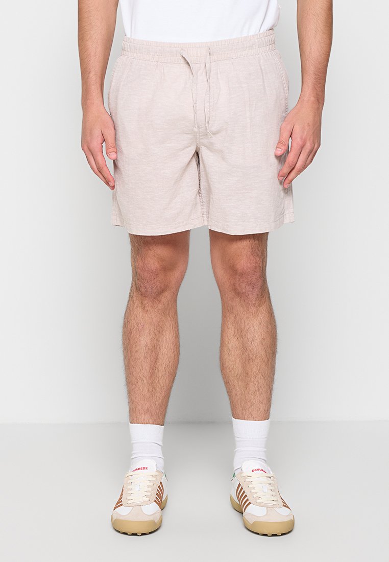 Homme portant un short beige clair à cordon, des chaussettes blanches et des baskets blanches avec des rayures beiges et marron, debout devant un fond uni.