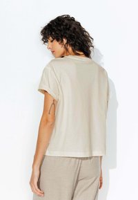T-shirt en coton beige avec des manches retroussées et une coupe décontractée, présentant un col rond et une texture lisse. Porté avec un pantalon gris clair.