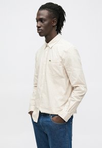 Bež in bela karo srajca z gumbi in zelenim logotipom, nošena z modrimi kavbojkami. Srajca ima klasičen ovratnik in dolge rokave.