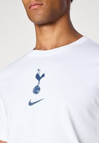Hvid t-shirt med et marineblåt grafisk motiv af en hane over en fodbold, med et lille Nike-logo nedenfor. Rund hals, korte ærmer.