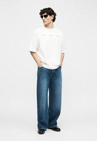 Άνδρας με μαύρα γυαλιά ηλίου, λευκό φαρδύ μπλουζάκι, φαρδιά μπλε τζιν και μαύρα παπούτσια, στέκεται με τα χέρια στις τσέπες μπροστά σε λευκό φόντο.