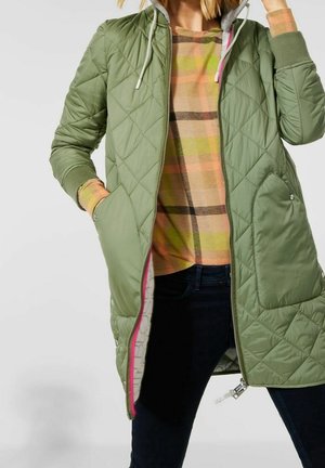 Femme portant une veste matelassée à capuche verte sur un pull pastel à carreaux et un pantalon foncé, une main dans la poche et l'autre ajustant la capuche.