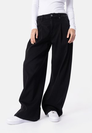 Persona che indossa pantaloni neri a gamba larga, una maglia bianca a maniche lunghe e scarpe da ginnastica bianche, in piedi davanti a uno sfondo semplice.