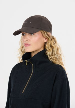 Kvinde med krøllet blondt hår iført en sort lynlåstrøje og en mørkebrun kasket med hvid teksten "ATHLECIA".