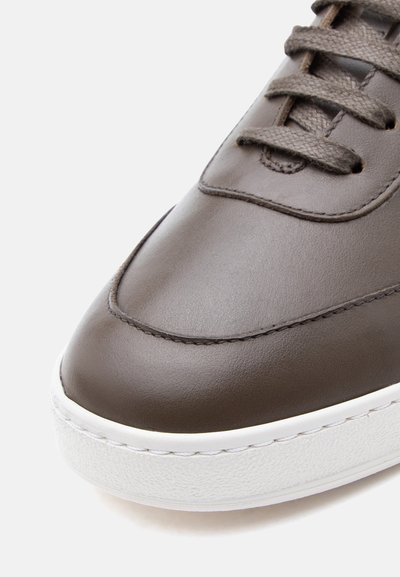 Καφέ δερμάτινο αθλητικό παπούτσι με υφασμένες κορδόνια και λευκή λαστιχένια σόλα, που αναδεικνύει την ομ αλή περιοχή στη μύτη και τις λεπτομέρειες ραφών.