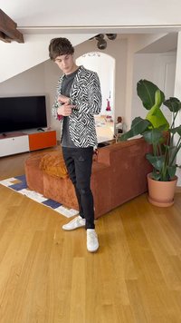 En sort-hvid zebra-stribet blazer over en grå skjorte, parret med slidte sorte bukser og hvide sneakers i en indendørs setting.
