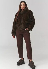 Brązowa krótka kurtka z futra sztucznego z wysokim kołnierzem, zestawiona z brązowymi spodniami i masywnymi czarnymi butami. Miękka tekstura, casualowy fason, przednie kieszenie.