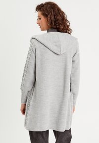 Lange grijze gebreide cardigan met een gestructureerde weefsel, oversized capuchon en gevlochten accenten op de mouwen. Zijslitsen verhogen de flexibiliteit.