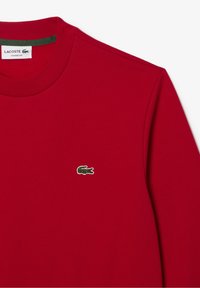 T-shirt rouge à manches longues en coton avec un col rond. Présente un petit logo Lacoste vert brodé sur la poitrine.