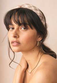 Ruudullinen kangaspanta beigen sävyissä, kierretty kullanvärinen kaulakoru ja pienet renkaankorvakorut; mallilla on aaltoilevat tummat hiukset ja sileä iho.