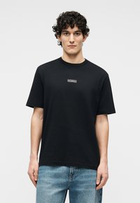 Hombre con cabello rizado vistiendo una camiseta negra de Iceberg y jeans azul claro, de pie contra un fondo blanco liso.
