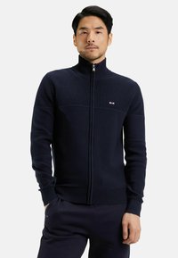 Sweat-shirt zippé marine avec texture côtelée, col haut et motif à rayures horizontales. Présente un petit logo sur la poitrine et des poignets ajustés.