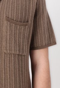 Cameră din tricot maro cu mânecă scurtă, cu un design texturat cu nervuri și detaliu de buzunar pe piept, realizată dintr-un material moale și ușor.
