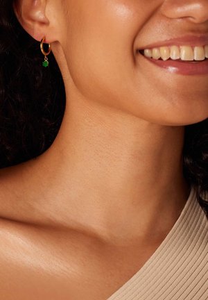 Gouden oorbel met een groene charme, gedragen aan het oor van een model. Krullend haar en een gladde huid zijn zichtbaar, samen met een getextureerde off-shoulder top.