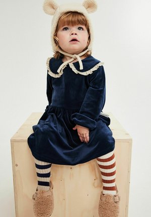 Granatowa aksamitna sukienka z falbanianym kołnierzem, zestawiona z paskowanymi rajstopami i futerkowymi brązowymi kapciami. Dziecko ma na głowie kremową czapkę z uszami misia.