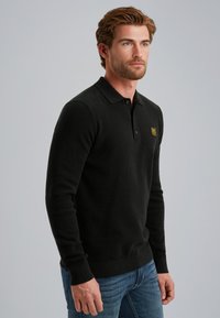 Zwarte langwerpige polo trui met een gestructureerde brei, voorzien van een kraag met knopen en een klein geel logo op de borst, gecombineerd met een blauwe spijkerbroek.