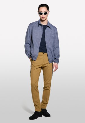 Veste texturée bleue, chemise marine, pantalon beige, et bottines noires. Mannequin portant des lunettes de soleil blanches, se tenant devant un fond clair.