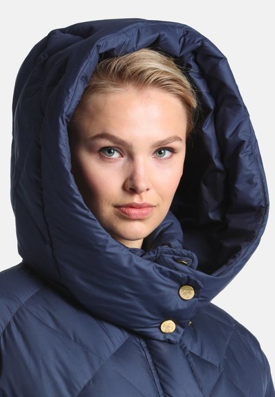 Femme aux cheveux clairs porte une veste matelassée bleu foncé avec une grande capuche bouffante et des boutons dorés, regardant droit devant elle.