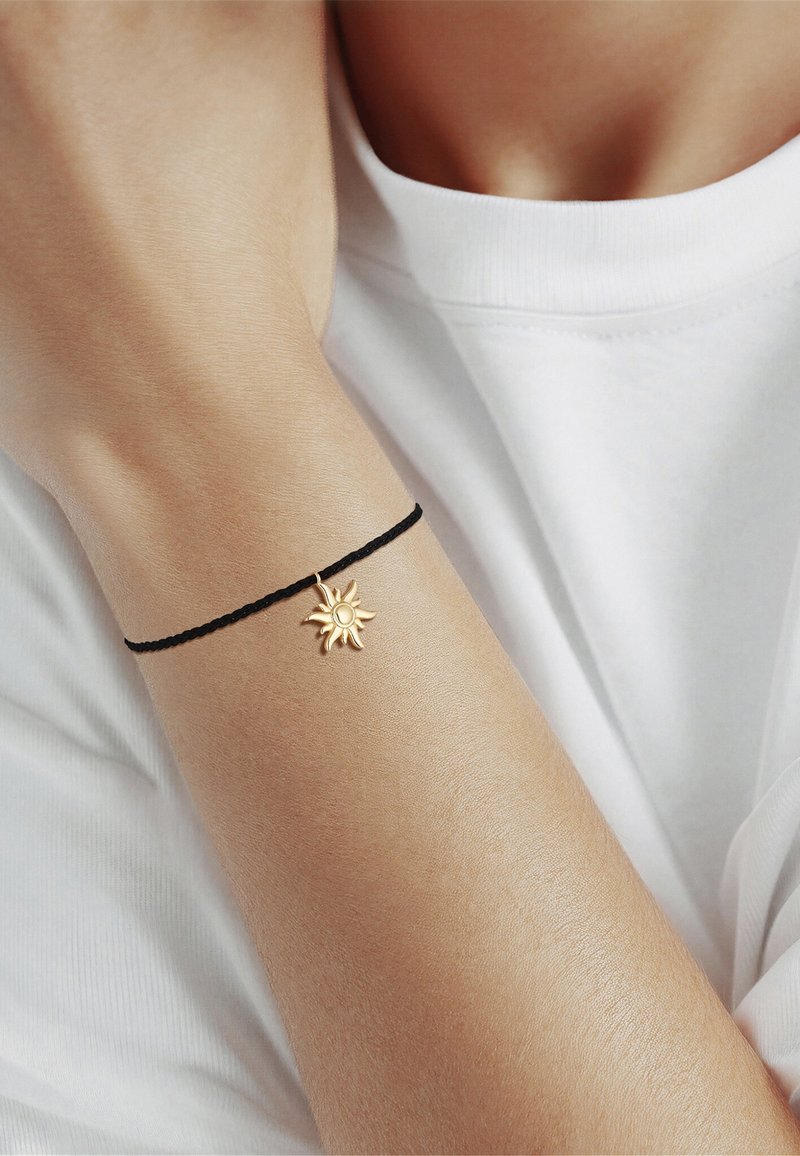 Arm med en tynd sort armbånd med en lille gylden solvedhæng, mod en hvid skjortebaggrund.