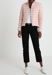 Person iført let lyserød quiltet jakke, hvid V-hals skjorte, sorte cropped bukser og hvide sneakers, stående foran en ensfarvet baggrnd.