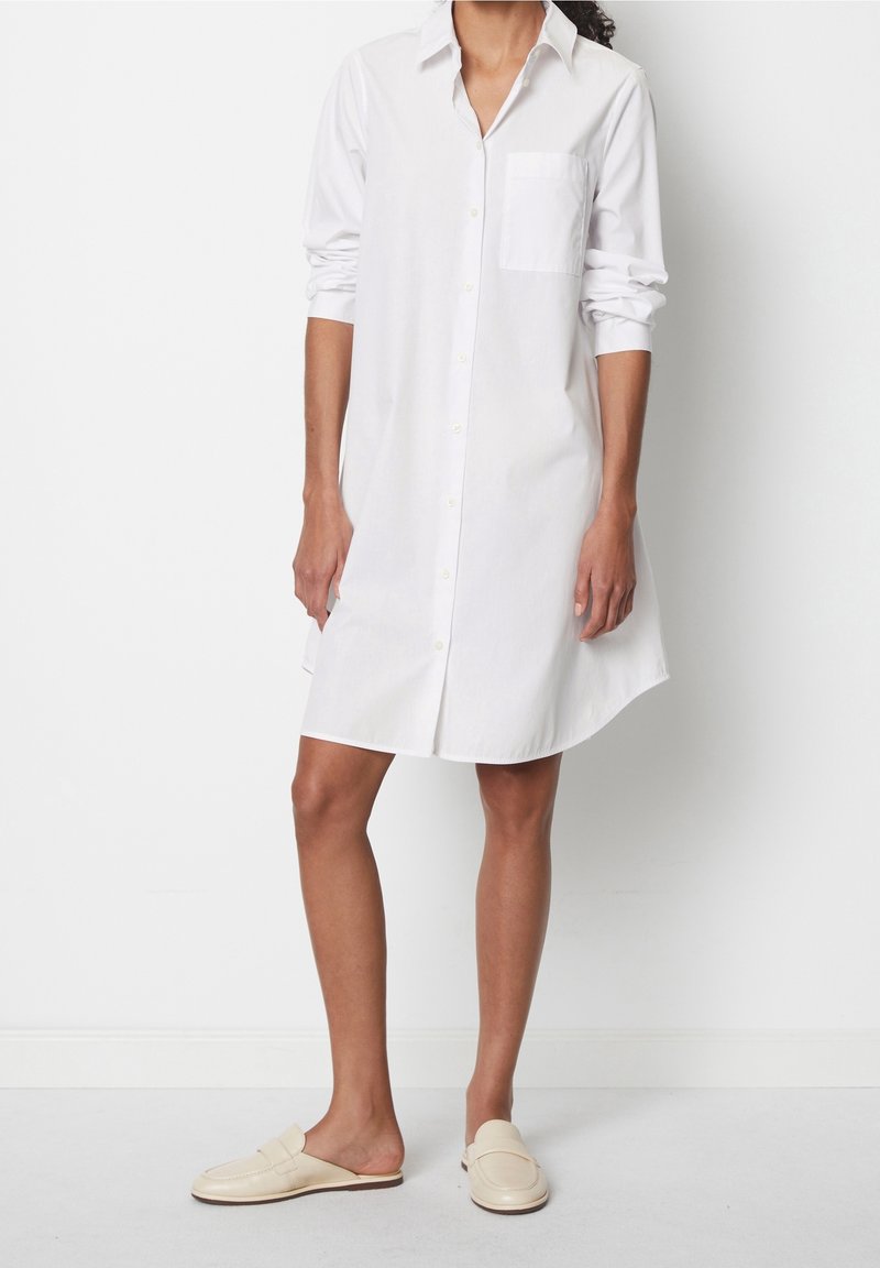 Femme portant une robe chemise blanche à manches longues boutonnée et des mocassins crème sans lacets, debout devant un fond blanc uni.