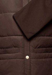 Veste matelassée marron avec surpiqûres horizontales, deux poches avant, un bouton-pression doré visible et un détail de poignet lisse.