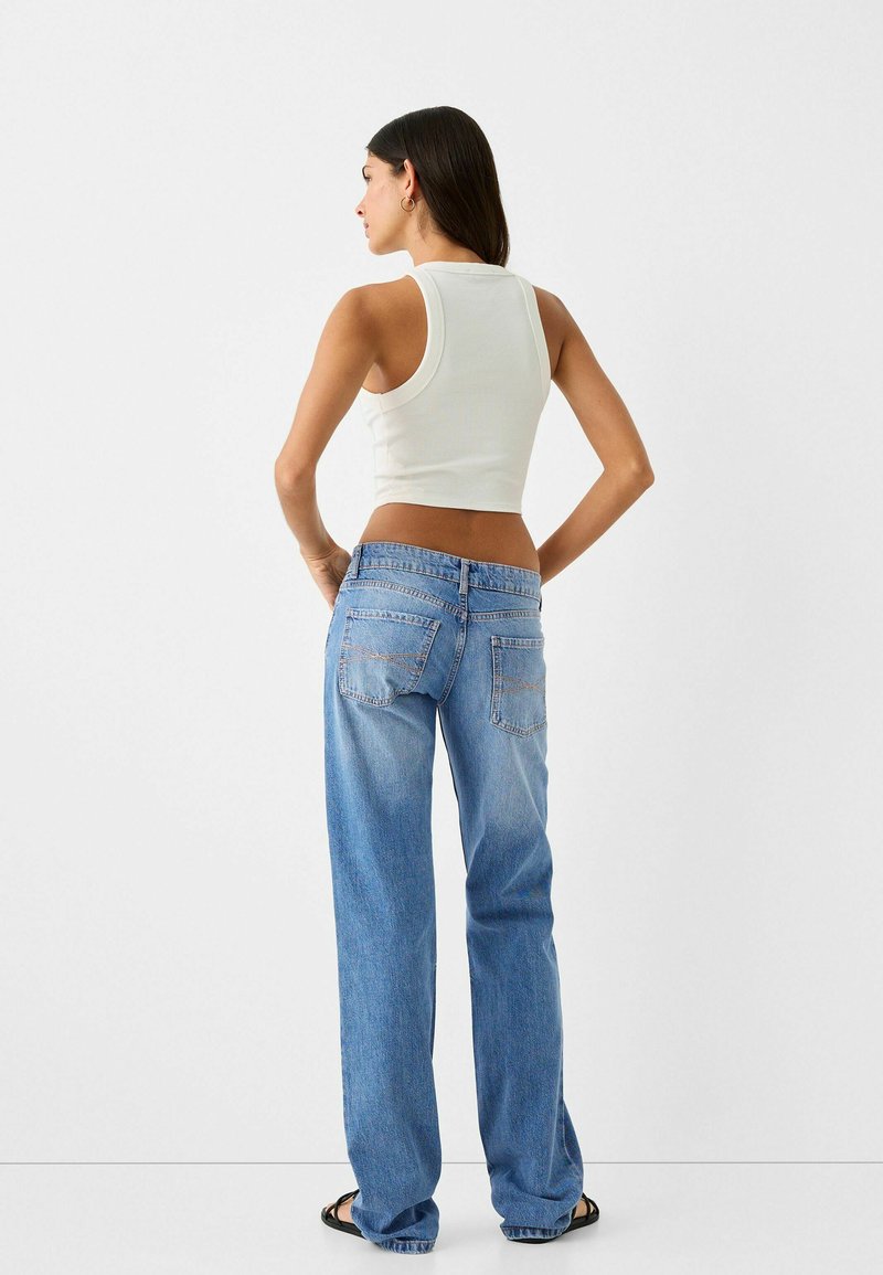 Top corto blanco, jeans sueltos de un azul claro con bolsillos traseros, manos del modelo en las caderas, fondo minimalista.