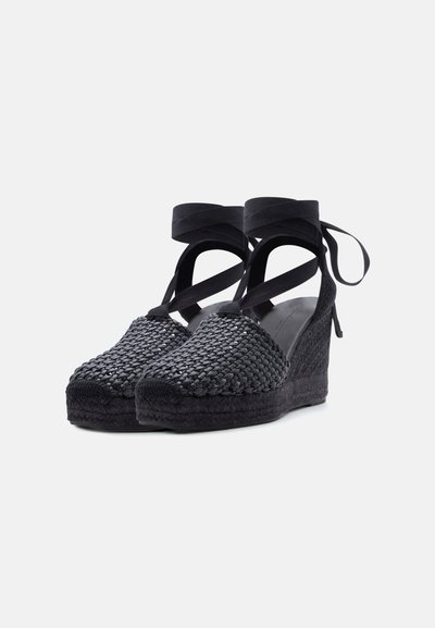 Espadrilles compensées noires tissées avec rubans à nouer autour de la cheville et bout fermé, présentées sur fond blanc.