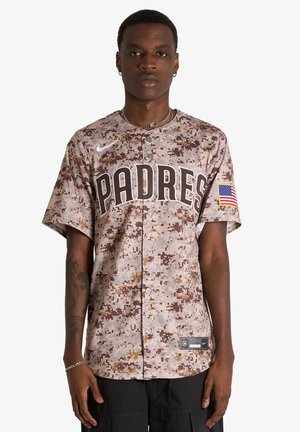 Giovane uomo che indossa una maglia da baseball dei Padres con motivo mimetico e una toppa della bandiera americana sulla manica e pantaloni neri.