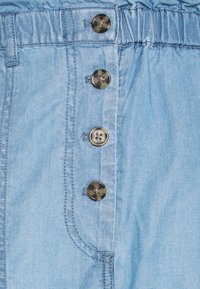 Tela de denim azul claro con un acabado brillante, que presenta cuatro botones oscuros, una cintura fruncida y costuras en contraste para un detalle adicional.