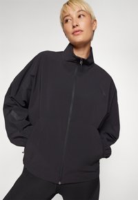 Black lightweight jacket with high collar, zip closure, and elastic cuffs. Features a smooth, matte finish and loose, relaxed fit.