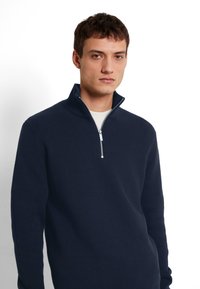 Marinblå stickad zip-up tröja med hög krage, med en silverfärgad dragkedja och ribbat mönster. Bärs över en ljusfärgad skjorta.