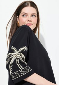 Top negro de corte relajado, con un bordado de palmeras en crema en la manga y un patrón de olas a lo largo del dobladillo. Textura de tela suave.