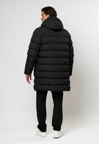 Mann som har på seg en lang svart polstret frakk med hette, svarte bukser og hvite sneakers, står vendt bort fra kameraet mot en ensfarget bakgrunn.