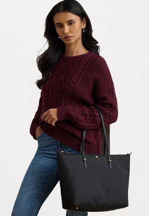 Bolsa tote preta feita de material liso, com duas alças longas em couro, detalhes em dourado e um logotipo na parte frontal.