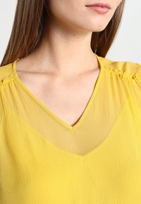 Žlutá, lehká blůza s výstřihem do V, nařasenými detaily na ramenou a měkkou, texturovanou látkou.