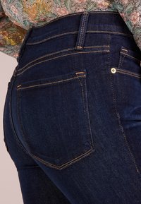 I jeans in denim blu scuro presentano un design a vita alta, cuciture dorate e due tasche posteriori con semplici cuciture orizzontali e rivetti.